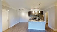 This single-floor residence showcases a thoughtfully designed open-concept layout that seamlessly connects the living and kitchen areas, creating a bright and inviting atmosphere. The kitchen is a standout feature, outfitted with rich dark cabinetry, granite countertops, and a full suite of stainless steel appliances, including a refrigerator, range, and microwave. A peninsula counter with pendant lighting serves as both a functional prep area and a casual dining space, adding an elegant focal point to the room. Track lighting runs across the ceiling in both the kitchen and living areas, ensuring consistent and flexible illumination throughout. The floors are finished with wood-style planking that adds warmth and continuity across the open space. Crown molding trims the ceilings, lending a refined architectural detail that elevates the overall aesthetic. The neutral color palette of soft whites and grays creates a clean, contemporary backdrop that feels both modern and welcoming. The open flow between the living and kitchen zones makes the space feel generous and well-suited for both everyday living and entertaining.