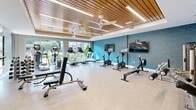 Located at 301 Continental Pkwy in Newport News, Virginia, this fitness center offers a thoughtfully designed exercise space across a single floor. The room's standout architectural feature is its warm wood-slat ceiling accented by recessed linear lighting and ceiling fans, creating a polished yet welcoming atmosphere. A vibrant teal textured accent wall adds a bold pop of color and serves as a visual focal point throughout the space.

Large floor-to-ceiling windows line one side of the room, flooding the interior with natural light and providing open views of the surrounding outdoor greenery. The light-toned, durable flooring is well-suited for an active environment and keeps the space feeling bright and clean.

The cardio zone is generously appointed with multiple treadmills, stationary bikes with individual digital screens, an elliptical trainer, and a rowing machine — all arranged to take advantage of the outdoor views. Wall-mounted televisions provide additional entertainment options. The strength and functional training area features weight benches, a cable machine, a dumbbell rack, kettlebells, and a full-length mirror wall that enhances the sense of openness. Resistance bands and other accessories are also available. Together, these elements create a comprehensive, modern fitness environment suited for a wide range of workout routines.