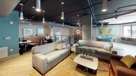 This expansive two-floor amenity facility is thoughtfully designed to serve a variety of social, recreational, and productive needs. The lower level opens into a vibrant lounge area anchored by a curved, wood-clad kitchen bar topped with a white countertop, illuminated by large industrial pendant lights against a bold teal accent wall. Adjacent seating areas feature cozy booth-style nooks and casual lounge furniture, ideal for working or gathering. A dedicated game room houses a full-size pool table, a shuffleboard table, and wall-mounted televisions, all beneath a dramatic dark ceiling with exposed ductwork and eclectic lighting fixtures. A bright co-working nook lined with a long counter and windows overlooking the building exterior provides a focused workspace. The upper level is accessed via a striking open staircase with reclaimed wood treads and a sleek steel-cable railing. Upstairs, an expansive lounge showcases clusters of contemporary sofas and chairs, artistic wall pieces, and abundant pendant lighting. A card table area with floor-to-ceiling glass doors opens to an outdoor courtyard view. Throughout both floors, light wood flooring, curated artwork, and a cohesive modern-industrial aesthetic create an inviting and energetic atmosphere.