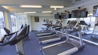 This fitness center is a dedicated single-floor space designed to support a comprehensive cardio and exercise routine. The room is outfitted with a lineup of modern cardio equipment, including multiple treadmills and elliptical machines arranged in an organized row, making efficient use of the available floor space. The equipment is manufactured by reputable brands such as Spirit and Life Fitness, reflecting a commitment to quality fitness resources.

Overhead, fluorescent lighting ensures the space is consistently and brightly illuminated throughout. Multiple flat-screen TVs are mounted on the walls, allowing users to stay entertained or informed during their workouts. Large windows along one wall invite natural light into the room and provide views of the surrounding exterior, adding an open and airy quality to the environment.

The flooring consists of durable, dark-toned carpet suited for a fitness environment, providing cushioning and stability underfoot. The walls are finished in a clean, neutral tone, contributing to a focused and energizing atmosphere. A sanitization station is also visible on the wall, emphasizing cleanliness and user convenience. Overall, this fitness center offers a practical, well-maintained environment suitable for daily exercise needs.