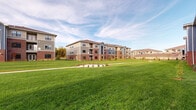 This residential community is composed of several three-story buildings arranged around generous stretches of well-maintained green lawn, creating an open, campus-like atmosphere. The architecture combines classic red brick bases with contemporary fiber cement siding in neutral and accent tones of gray and blue, capped by pitched roofs with dark shingles. This blend of traditional and modern materials gives the exterior a durable yet visually appealing character.

Each building features large, dark-framed windows that allow ample natural light, while select units include private balconies enclosed by black metal railings—offering outdoor space with views overlooking the surrounding grounds. Ground-level units benefit from covered patios with matching railings and direct access to landscaped planting beds filled with mulch and low shrubs.

The shared outdoor spaces are a standout feature of the community, with broad expanses of lush green lawn connecting the buildings. Concrete pedestrian pathways wind through the grounds, and newly planted trees suggest continued landscape development. The open sightlines across the property create a sense of spaciousness and connectivity between buildings, contributing to an inviting, well-planned community environment.