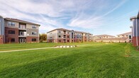 This residential community is composed of several three-story buildings arranged around generous stretches of well-maintained green lawn, creating an open, campus-like atmosphere. The architecture combines classic red brick bases with contemporary fiber cement siding in neutral and accent tones of gray and blue, capped by pitched roofs with dark shingles. This blend of traditional and modern materials gives the exterior a durable yet visually appealing character.

Each building features large, dark-framed windows that allow ample natural light, while select units include private balconies enclosed by black metal railings—offering outdoor space with views overlooking the surrounding grounds. Ground-level units benefit from covered patios with matching railings and direct access to landscaped planting beds filled with mulch and low shrubs.

The shared outdoor spaces are a standout feature of the community, with broad expanses of lush green lawn connecting the buildings. Concrete pedestrian pathways wind through the grounds, and newly planted trees suggest continued landscape development. The open sightlines across the property create a sense of spaciousness and connectivity between buildings, contributing to an inviting, well-planned community environment.