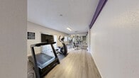 Located at 1900 East Tropicana Avenue in Las Vegas, NV, this fitness center offers a practical and well-equipped exercise space designed to support a range of workout routines. The room features light-toned, wood-look flooring that is both durable and easy to maintain, complemented by neutral gray walls and a textured ceiling with flush lighting that keeps the space bright and functional.

Cardio equipment includes a treadmill with an integrated display monitor and an elliptical trainer, both positioned to make efficient use of the available floor space. A multi-station cable weight machine with an adjustable bench provides options for a variety of strength training exercises. Full-length mirrors line one wall, enhancing the sense of space and allowing users to monitor their form during workouts.

Natural light enters through windows fitted with vertical blinds, offering both illumination and privacy control. A purple accent detail near the ceiling adds a subtle touch of branding or color contrast to the otherwise clean, minimalist interior. The layout is straightforward and efficiently organized, making the most of the compact footprint. Two access doors provide convenient entry and exit points. Overall, the fitness center delivers a focused, no-frills workout environment suited for daily use by residents or guests of the surrounding property.