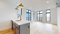 This contemporary single-floor unit showcases a thoughtfully designed open-concept layout that seamlessly connects the kitchen and living areas. Light natural hardwood flooring runs throughout, lending warmth and continuity to every room. Large black-framed windows are a defining architectural feature, flooding the interior with abundant natural light while offering views of the surrounding neighborhood. Several windows are accompanied by sleek metal balcony railings, adding an industrial-modern aesthetic.

The kitchen is a standout space, centered around a substantial island with a crisp white quartz countertop, an undermount sink, and a matte black faucet. Bold blue-grey cabinetry pairs with stainless steel appliances, including a refrigerator, range, and microwave. Pendant lighting above the island adds both function and visual interest.

The bedrooms are bright and spacious, each benefiting from the generous window placement and ceiling fans for comfort. Exposed silver ductwork in select rooms adds an industrial touch that complements the modern design language.

The bathrooms are elegantly finished with dark hexagonal floor tiles, white subway tile walls, matte black fixtures, and arched mirrors, creating a clean and sophisticated aesthetic. One bathroom features a walk-in shower, while another includes a full soaking tub with an open glass partition.