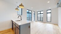 This contemporary single-floor unit showcases a thoughtfully designed open-concept layout that seamlessly connects the kitchen and living areas. Light natural hardwood flooring runs throughout, lending warmth and continuity to every room. Large black-framed windows are a defining architectural feature, flooding the interior with abundant natural light while offering views of the surrounding neighborhood. Several windows are accompanied by sleek metal balcony railings, adding an industrial-modern aesthetic.

The kitchen is a standout space, centered around a substantial island with a crisp white quartz countertop, an undermount sink, and a matte black faucet. Bold blue-grey cabinetry pairs with stainless steel appliances, including a refrigerator, range, and microwave. Pendant lighting above the island adds both function and visual interest.

The bedrooms are bright and spacious, each benefiting from the generous window placement and ceiling fans for comfort. Exposed silver ductwork in select rooms adds an industrial touch that complements the modern design language.

The bathrooms are elegantly finished with dark hexagonal floor tiles, white subway tile walls, matte black fixtures, and arched mirrors, creating a clean and sophisticated aesthetic. One bathroom features a walk-in shower, while another includes a full soaking tub with an open glass partition.