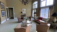 This single-floor space makes a striking first impression with its dramatic double-height ceilings and expansive arched windows that flood the interior with natural light. The main living area is generously proportioned, offering a sense of openness and grandeur that is both welcoming and refined.

The living room serves as the centerpiece of the space, anchored by a decorative wrought-iron chandelier that adds an elegant, artistic focal point overhead. Warm taupe-toned walls provide a neutral backdrop that complements the richly patterned upholstered seating, including floral armchairs and a neutral sofa accented with deep red throw pillows. A metal and glass coffee table sits at the heart of the seating arrangement, topped with a lush potted plant that adds a touch of organic warmth.

Large floor-to-ceiling windows with draped curtains frame views of an outdoor deck and surrounding greenery, seamlessly connecting the interior to the natural landscape beyond. The light-toned flooring runs throughout, contributing to the airy, open feel of the space.

Thoughtful touches such as accent lamps, framed wall art, and carefully arranged decor give the space a curated, lived-in charm. Overall, the space balances classic architectural details with comfortable furnishings to create a sophisticated yet approachable environment.