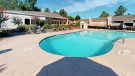 Located at 2525 Center West Pkwy in Augusta, Georgia, this outdoor amenity area is designed for relaxation and recreation. At its centerpiece is a generously sized, kidney-shaped swimming pool with crystal-clear turquoise water, accessible via stainless steel entry rails and surrounded by an expansive stamped concrete sun deck. The deck accommodates multiple lounge chairs and seating areas, ideal for sunbathing and socializing. A dedicated patio seating area features a round table with chairs shaded by a large umbrella, offering a comfortable spot to unwind. Adjacent to the pool deck, a built-in stone fire pit is surrounded by additional lounge seating, creating an inviting gathering space for cooler evenings. The entire pool area is enclosed by a black wrought-iron fence, ensuring security while maintaining open sightlines to the lush, landscaped grounds. Mature trees, blooming crape myrtles, and well-maintained shrubs frame the perimeter, adding vibrant color and natural beauty to the setting. Residential buildings are visible in the background, nestled among the greenery and set beneath open Georgia skies, contributing to the welcoming, community-oriented atmosphere of this outdoor space.