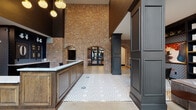 This single-floor lobby space makes an immediate architectural statement through its dramatic, soaring ceiling height and expansive full-height glazed entry doors that flood the interior with natural light. The dominant design feature is the full-height exposed brick wall, punctuated by arched brick openings that frame views into adjoining areas, lending the space a historic industrial character balanced by refined modern finishes.

The flooring transitions between polished concrete and an eye-catching black-and-white geometric patterned tile that anchors the central circulation zone. A substantial reception desk crafted from warm wood paneling with a crisp white countertop serves as a functional focal point, backed by a dark built-in display unit featuring decorative accessories and a vintage-style number board.

Clustered globe pendant lighting and an angular metallic wall sculpture add artistic dimension to the dark charcoal-painted upper walls. Structural columns with paneled detailing define separate zones within the open plan, including a comfortable lounge area with accent chairs and a dedicated library nook featuring floor-to-ceiling dark built-in shelving, a solid wood communal table, and plush seating. Together, these elements create a sophisticated, boutique-style atmosphere.