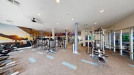 Located at 1660 Metro Ave in Chula Vista, this fitness studio offers a generously sized, single-floor workout facility designed to accommodate a full spectrum of exercise needs. The open-plan layout is anchored by light-colored carpet with teal accent tiles, giving the space a clean, modern aesthetic. Structural columns with blue accents are distributed throughout the floor, supporting a bright white ceiling fitted with recessed lighting and ceiling fans for optimal comfort and ventilation.

The strength training area is well-appointed with an extensive selection of selectorized machines, cable systems, free-weight benches, and multiple racks of dumbbells. Full-length mirrors along the walls enhance the sense of space and support proper form during workouts. The cardio section features a row of elliptical trainers and stationary bikes positioned alongside large floor-to-ceiling windows, flooding the area with natural light and offering views of the surrounding outdoor landscaping.

A glass-enclosed room provides a dedicated space for specialized training or classes. Vibrant, large-scale motivational murals depicting athletes and wellness imagery are displayed throughout, adding energy and visual interest to the environment. The studio's spacious, well-organized layout ensures ample room between equipment, creating a comfortable and functional workout experience.