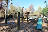 This outdoor recreational space is nestled within a mature, wooded setting that provides a naturally serene atmosphere throughout. A network of winding concrete pathways connects the various amenities, weaving between tall trees whose canopy offers shade and a sense of seclusion from the surrounding area.

A standout feature is the large wooden pavilion, constructed with an exposed timber-framed roof and open-air design. Enclosed by wooden railings, the pavilion provides a shaded gathering area furnished with seating and tables, making it well-suited for group activities or relaxed outdoor enjoyment.

Adjacent to the pavilion, a full-size tennis court is enclosed by a chain-link fence and accessed via a wooden deck equipped with railings and posted regulations. The court surface appears well-maintained and benefits from the open sky above.

A playground area is also integrated into the space, featuring a swing set, a slide structure, and additional play equipment set on a ground cover of natural materials. Trash receptacles are conveniently placed nearby along the pathways.

The overall ambiance is defined by the dense tree coverage, seasonal foliage, and natural ground cover, creating a peaceful, park-like environment that encourages outdoor activity and community engagement.