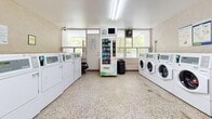 Located at 45 Balliol St in Toronto's Davisville Village neighbourhood, this shared laundry facility offers a well-equipped, functional space designed to serve a residential building. The room is illuminated by overhead fluorescent lighting and benefits from generous natural light streaming through large windows that overlook mature trees outside, creating a bright and open atmosphere.

Rows of front-loading washing machines and top-loading dryers line opposing walls, providing ample capacity for multiple users simultaneously. A dedicated utility sink with cabinetry is conveniently situated along one wall, offering space for hand-washing or pre-treating garments. A centrally positioned vending machine stocks laundry supplies, adding day-to-day convenience for residents.

The floor throughout is finished in durable, speckled terrazzo, a practical and long-lasting surface well-suited to high-traffic environments. The walls feature a neutral tile and painted finish, maintaining a clean and tidy appearance. A card-operated payment kiosk is available on-site, streamlining the payment process. Safety features include a fire extinguisher and clearly posted notices. Multiple entry and exit points provide easy access, and the overall layout ensures comfortable circulation throughout the space.