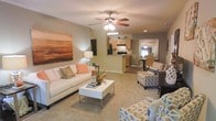 Located at 2850 Courtney Creek Blvd in Durham, North Carolina, this single-floor residence offers a well-designed layout that balances comfort and functionality. The open-concept living area serves as the heart of the home, featuring plush carpeting, a ceiling fan with integrated lighting, and recessed lights that provide warm, even illumination throughout. Large-scale wall art adds a bold visual anchor to the living space, which is generously sized to accommodate multiple seating arrangements. The neutral color palette of soft beige and warm gray tones creates a welcoming and versatile atmosphere. The living area flows naturally into a kitchen that features wooden cabinetry, dark appliances, and a breakfast bar-style counter that adds a casual dining option. Adjacent to the kitchen, a dedicated dining area with seating for multiple guests is visible through an arched passageway, enhancing the sense of connected, functional living. The hardwood-style flooring in the kitchen and dining areas provides a clean contrast to the carpeted living space. The overall design emphasizes an open, airy feel with a cohesive layout that maximizes the usability of every area within the home.