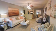 Located at 2850 Courtney Creek Blvd in Durham, North Carolina, this single-floor residence offers a well-designed layout that balances comfort and functionality. The open-concept living area serves as the heart of the home, featuring plush carpeting, a ceiling fan with integrated lighting, and recessed lights that provide warm, even illumination throughout. Large-scale wall art adds a bold visual anchor to the living space, which is generously sized to accommodate multiple seating arrangements. The neutral color palette of soft beige and warm gray tones creates a welcoming and versatile atmosphere. The living area flows naturally into a kitchen that features wooden cabinetry, dark appliances, and a breakfast bar-style counter that adds a casual dining option. Adjacent to the kitchen, a dedicated dining area with seating for multiple guests is visible through an arched passageway, enhancing the sense of connected, functional living. The hardwood-style flooring in the kitchen and dining areas provides a clean contrast to the carpeted living space. The overall design emphasizes an open, airy feel with a cohesive layout that maximizes the usability of every area within the home.