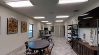 This single-floor space offers a welcoming and functional environment designed for casual gatherings, meetings, or social use. The main area is outfitted with multiple round dark-topped tables paired with industrial-style metal chairs, creating a relaxed and versatile seating arrangement that accommodates small groups comfortably.

The room features light-toned tile flooring with a wood-look pattern, white walls, and a neutral color palette that contributes to a clean and open atmosphere. Overhead, flush-mounted fluorescent light fixtures provide consistent, even illumination throughout the space.

Along one wall, a metal shelving unit holds decorative accents, books, and various objects, adding character to the room. A small service counter or bar area is visible toward the back of the space, accompanied by additional seating — suggesting a refreshment or check-in station. Decorative wall art, including a framed playing card graphic, adds a touch of personality to the interior.

Toward the rear of the room, two interior doors lead to additional spaces, likely restrooms or utility areas. Windows fitted with horizontal blinds allow natural light to filter in while maintaining privacy. The overall design is practical and well-organized, balancing functional layout with modest decorative elements suited for a community or multi-purpose facility.