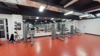 This fitness center is designed to support a full range of workout activities within a single, open-plan floor. The space features bold red rubberized flooring throughout, providing both durability and a dynamic aesthetic that contrasts with the dark-painted exposed ceiling overhead. Industrial-style HVAC ductwork and overhead fluorescent lighting run across the ceiling, delivering consistent, bright illumination across the entire facility.

Large floor-to-ceiling mirrors line multiple walls, visually expanding the space and allowing members to monitor their form during exercises. The cardio zone is well-stocked with a row of treadmills and elliptical machines, each equipped with individual consoles, arranged for easy access and spacing. Wall-mounted flat-screen televisions are positioned for visibility throughout the cardio area.

The strength-training section houses a variety of cable and resistance machines, including chest press, lat pulldown, and multi-functional cable stations. A dedicated free weights area includes a full rack of dumbbells in graduated sizes, stability balls, resistance bands, and ankle weights. Vending machines near the perimeter offer convenient access to refreshments and snacks. The layout is open and functional, providing ample floor space for movement between equipment zones.
