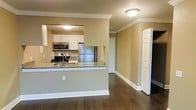 This single-floor residence offers a thoughtfully designed open-concept layout that seamlessly connects the kitchen and living areas. The kitchen is a standout feature, equipped with stainless steel appliances — including a refrigerator, range, and built-in microwave — complemented by white cabinetry and granite countertops. A peninsula with a granite overhang serves as a natural divider between the kitchen and the adjacent living space, providing both additional prep space and a casual seating area. Rich, dark hardwood flooring extends throughout the main living area, adding warmth and contrast to the neutral, tan-toned walls. Elegant crown molding runs along the ceilings, lending a refined, classic character to the interior. Flush-mount ceiling lights provide consistent, ambient illumination throughout. Crisp white baseboards and trim work provide clean definition along the walls and doorways. The overall ambiance is warm, clean, and move-in ready, with a balance of modern appliances and traditional architectural detailing that creates a welcoming and functional living environment.