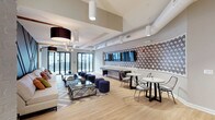 This thoughtfully designed clubhouse offers a rich blend of contemporary style and functional amenity spaces across a single, expansive floor. The main lounge area immediately draws attention with its striking exposed dark wood ceiling beams, light hardwood flooring, and a curved sofa arrangement anchored by a bold yellow-green accent partition — creating an inviting focal point for social gatherings. White painted brick columns and walls add an industrial-chic texture that contrasts beautifully with the refined furnishings.

A dedicated media lounge features multiple wall-mounted flat-screen televisions set against a dramatic dark accent wall with decorative tile detailing, flanked by deep sectional sofas and ottomans for comfortable viewing. Adjacent to this, a kitchen and bar area showcases elegant marble-style countertops, a statement chandelier, and direct access to an outdoor terrace through large glass doors, allowing natural light to pour in.

Private work nooks with built-in white desks, stylish sconce lighting, and patterned carpet offer quiet spaces for individual focus. A dedicated conference room with a round table, oversized pendant light, and a mounted display screen provides a professional meeting environment. Throughout, the clubhouse balances communal energy with intimate spaces, making it a versatile and visually compelling amenity.