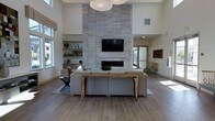 This single-floor space showcases a striking contemporary design anchored by a dramatic floor-to-ceiling tiled fireplace wall in soft grey tones. A wall-mounted flat-screen television is integrated seamlessly above the linear fireplace, creating a bold and functional focal point. The living area is generously proportioned, with soaring double-height ceilings that amplify the sense of openness and airiness throughout the space.

Wide-plank wood-style flooring in warm, neutral tones runs throughout, complementing the modern yet inviting aesthetic. A large sectional sofa in soft grey is paired with a live-edge wood console table, adding an organic element that balances the clean architectural lines. Built-in open shelving to the side of the fireplace provides display space and adds architectural depth.

Natural light pours in through large windows dressed with roman shades, illuminating the space without overwhelming it. An oversized modern pendant light fixture anchors the ceiling above the seating area. A secondary seating nook is visible toward the rear, hinting at a versatile, multi-use layout. The overall ambiance is calm, refined, and welcoming.