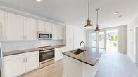 Located at 3612 Beverly Cove Way in Wilmington, NC, this thoughtfully designed one-bedroom, one-bathroom residence offers a clean, modern aesthetic highlighted by light wood-look flooring and crisp white trim throughout. The open-concept kitchen serves as the centerpiece of the home, featuring white shaker-style cabinetry, a classic subway tile backsplash, dark quartz countertops, and a full suite of stainless steel appliances including a refrigerator with ice maker, electric range, and built-in microwave. A central kitchen island with an undermount sink and bronze pendant lighting above adds both function and style, flowing seamlessly into the living area. French doors at the rear of the living space invite natural light and provide outdoor access. The bedroom is comfortably proportioned, outfitted with crown molding, a ceiling fan, and dual windows offering views of the surrounding community. A generously sized walk-in closet with wire shelving provides ample storage. The bathroom is finished with a white vanity with dark countertop, a tall linen cabinet, gold-toned fixtures, and wood-look flooring that ties the space together cohesively. Crown molding and recessed lighting are present throughout, lending a polished, refined character to this efficiently designed home.