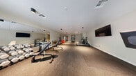 Located at 1100 D St in Las Vegas, Nevada, this fitness center offers a generously sized, single-floor exercise room designed to accommodate a full range of workout activities. The space features durable speckled rubber flooring throughout, providing a safe and resilient surface ideal for both cardio and strength training. The clean, modern interior is finished with neutral-toned walls and a bright white ceiling equipped with recessed and surface-mounted lighting fixtures that ensure the room is well-illuminated at all times. A large wall-to-wall mirror on one side of the room enhances the sense of space and allows users to monitor their form during exercise. The facility is equipped with a variety of cardio machines, including treadmills, an elliptical trainer, and a stationary bike, as well as a dedicated strength-training area featuring a power rack, adjustable bench, and a full dumbbell rack with an extensive weight selection. A wall-mounted flat-screen television adds to the overall experience. Double glass doors at one end of the room invite natural light into the space and provide convenient access. An adjacent frosted-glass sliding door leads to a secondary area, further adding to the facility's functionality. The overall ambiance is clean, professional, and purpose-built for fitness.
