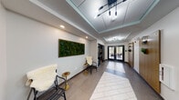 This contemporary single-floor commercial space is designed to make a strong first impression through its thoughtfully appointed entrance corridor. The hallway serves as the architectural spine of the space, featuring a striking herringbone-patterned tile inlay set against large-format neutral floor tiles, guiding visitors through the interior with visual purpose.

Overhead, recessed coffers with teal-accented drop ceilings add architectural depth, while a series of modern multi-bulb pendant and chandelier-style light fixtures provide warm, layered illumination throughout. Wall-mounted sconces with an industrial-modern aesthetic complement the overall lighting design and contribute to a polished ambiance.

A standout decorative element is the large framed living moss wall panel, which introduces a vibrant natural accent against the crisp white walls and adds a biophilic touch to the professional environment. Seating alcoves along the corridor feature contemporary accent chairs and gold-toned side tables, offering welcoming rest areas for visitors.

Black-framed glass double doors serve as both functional entry points and elegant visual dividers, allowing natural light to flow through the space and providing views into adjacent rooms. Glass partitions reveal well-lit office and meeting spaces. Warm wood-toned doors and wall-mounted greenery add organic warmth, balancing the clean modern aesthetic throughout this refined and functional commercial interior.