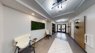 This contemporary single-floor commercial space is designed to make a strong first impression through its thoughtfully appointed entrance corridor. The hallway serves as the architectural spine of the space, featuring a striking herringbone-patterned tile inlay set against large-format neutral floor tiles, guiding visitors through the interior with visual purpose.

Overhead, recessed coffers with teal-accented drop ceilings add architectural depth, while a series of modern multi-bulb pendant and chandelier-style light fixtures provide warm, layered illumination throughout. Wall-mounted sconces with an industrial-modern aesthetic complement the overall lighting design and contribute to a polished ambiance.

A standout decorative element is the large framed living moss wall panel, which introduces a vibrant natural accent against the crisp white walls and adds a biophilic touch to the professional environment. Seating alcoves along the corridor feature contemporary accent chairs and gold-toned side tables, offering welcoming rest areas for visitors.

Black-framed glass double doors serve as both functional entry points and elegant visual dividers, allowing natural light to flow through the space and providing views into adjacent rooms. Glass partitions reveal well-lit office and meeting spaces. Warm wood-toned doors and wall-mounted greenery add organic warmth, balancing the clean modern aesthetic throughout this refined and functional commercial interior.