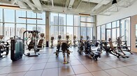 This contemporary fitness center occupies an elevated floor of a high-rise building, delivering an exceptional workout environment defined by soaring ceilings, industrial-chic exposed structural elements, and sweeping panoramic views of a dynamic urban skyline. Floor-to-ceiling curtain-wall glazing lines the perimeter, flooding the entire space with abundant natural light throughout the day.

The cardio zone is extensively equipped with a full lineup of ellipticals, stair climbers, stationary bikes, and treadmills—all positioned directly facing the city views, making every workout visually engaging. A dedicated strength training area features a comprehensive selection of plate-loaded machines, cable systems, and free weights, illuminated by striking linear pendant light fixtures suspended from the exposed concrete ceiling above.

The fitness center also includes generously sized open-floor studio spaces finished with light wood-tone flooring, pendant globe lighting, and glass-partitioned walls that maintain visual connectivity throughout. A wooden barre rail runs along one of the floor-to-ceiling window walls, suggesting dedicated space for yoga, stretching, or group fitness classes. A vibrant mural adds an artistic focal point to one section of the facility. The combination of industrial architecture, premium equipment, and unobstructed city vistas creates a motivating and sophisticated fitness environment.