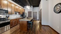 Located on St. Germain Street West in Saint Cloud, MN, this 1-bedroom, 2-bathroom unit showcases a striking industrial-chic aesthetic throughout. Dramatic exposed black ceilings with structural beams and track lighting create impressive vertical volume, complemented by rich, dark hardwood floors that flow seamlessly through the open-concept main living area.

The kitchen is a standout feature, offering warm medium-toned wood cabinetry extending to ceiling height, granite countertops, stainless steel appliances including a French door refrigerator and dishwasher, and a generous central island with bar seating — ideal for both cooking and casual dining. The kitchen flows directly into the living area, which benefits from a large sliding glass door that opens to a private balcony overlooking the streetscape, filling the space with natural light.

The bedroom provides a comfortable retreat with carpeted flooring, an exposed ceiling detail, and a window with urban views. A spacious walk-in closet with built-in shelving offers generous storage. Both bathrooms are cleanly finished, with one featuring a full tub-shower combination, tile flooring, and warm wood vanity cabinetry. The overall ambiance is modern and urban, blending industrial architecture with warm, residential finishes throughout.
