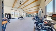 Located at 10670 Jake Jabs Blvd in Firestone, Colorado, this fitness center is a thoughtfully designed, generously proportioned facility that offers a comprehensive workout environment. The space is divided into two distinct zones: a main gym area with vaulted ceilings accented by exposed wood beams and dual ceiling fans, and a separate functional training room with light hardwood flooring and floor-to-ceiling windows overlooking an outdoor pool area.

The main gym area features neutral-toned carpet flooring and is bathed in natural light from large windows fitted with roller shades. A striking artistic mural depicting an abstract mountain landscape anchors one wall, adding a Colorado-inspired visual element to the space. Full-length mirrors amplify the sense of openness. Equipment includes Freemotion and CoachBike spin bikes, treadmills, ellipticals, a stair climber, rowing machines, a cable/power rack system, adjustable benches, and a dumbbell rack.

The functional training annex offers hardwood flooring and houses a FlaX functional training rack stocked with kettlebells, medicine balls, resistance bands, and battle ropes, along with plyo boxes and stability balls. Recessed lighting throughout both areas ensures a bright, welcoming atmosphere. The overall design strikes a balance between modern aesthetics and practical functionality, creating a motivating environment for a wide range of fitness activities.