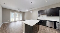 Located at 5005 Galleria Dr in Farmers Branch, TX, this thoughtfully designed one-bedroom, one-bathroom residence offers a comfortable and modern living experience across a single floor. The open-concept kitchen and living area serves as the heart of the home, showcasing rich dark cabinetry, white quartz countertops, a large central island, and stainless steel appliances including a refrigerator, range, microwave, and dishwasher. A tile backsplash and pendant lighting add refined touches to the kitchen. Wood-style plank flooring flows throughout the main living areas, complementing the warm, neutral wall tones. The living room is bright and airy, with French doors that invite natural light and provide outdoor access, further enhanced by a ceiling fan overhead. The spacious bedroom features plush carpeting, multiple windows with natural light, elevated ceilings, a ceiling fan, and direct access to both the bathroom and a walk-in closet with built-in shelving. The bathroom is finished with tile flooring, a dark vanity with quartz countertops, and a tub-shower combination. A hallway connects the private spaces seamlessly, rounding out this efficiently designed and well-finished residence.