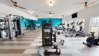 Located at 4800 N Star Ave in Panama City, Florida, this well-equipped fitness center offers a comprehensive workout environment designed to support a variety of exercise routines. The single-floor facility is spacious and open, with high ceilings and multiple ceiling fans that contribute to a comfortable, well-ventilated atmosphere. A bold teal accent wall energizes the space and provides a vibrant visual focal point, complemented by crisp white walls and a distinctive striped carpet tile flooring throughout.

The cardio zone features a lineup of treadmills, elliptical trainers, and stationary bikes, including interactive cycling equipment with digital displays. Ample natural light filters through several windows along the perimeter, creating a bright and motivating environment. Mounted flat-screen TVs offer additional entertainment options during workouts.

The strength training area is equally well-appointed, with a multi-station cable machine, a functional training rack stocked with resistance bands, medicine balls, and plyo boxes, as well as a dumbbell rack and weight bench. Full-length mirrors are positioned along the walls to assist with form and technique. The thoughtful layout ensures generous circulation space between equipment, making the facility both functional and comfortable for users of all fitness levels.