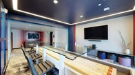 This contemporary single-floor space makes an immediate impression with its striking design aesthetic. The most prominent architectural feature is the deep navy-blue ceiling, accented by sleek LED strip lighting that runs along its perimeter, casting a warm, ambient glow throughout the room and lending a sophisticated, upscale atmosphere.

The space is thoughtfully divided into distinct functional zones. A central bar or counter area features a glass-topped surface with light wood paneling, flanked by cushioned seating with charcoal gray upholstery — ideal for casual gatherings or social interaction. Adjacent to this, a media lounge area is anchored by a large wall-mounted flat-screen television paired with a soundbar, set against clean white walls for a polished contrast.

The flooring features a patterned carpet that adds texture and visual interest underfoot, while white structural columns and red accent panels contribute bold, graphic elements to the overall design. In the background, a framed artwork piece with vivid blue tones adds a creative flourish. Recessed ceiling lighting supplements the LED accents, ensuring the space is well-lit. The open layout and varied seating options make this a versatile and inviting communal environment.