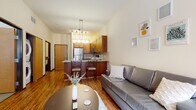 This single-floor unit offers a well-organized, open-concept layout that seamlessly connects the living, dining, and kitchen areas. The living space features warm hardwood-style flooring, a gray leather sofa, and a round coffee table, anchored by a large window that brings in natural light and urban views of the surrounding cityscape. Exposed ductwork near the window adds an industrial design accent.

The kitchen is efficiently designed with warm honey-toned cabinetry, a tile backsplash, and a full suite of stainless steel appliances including a refrigerator, range, and microwave. A peninsula with a granite-style countertop and bar seating for two provides both additional prep space and casual dining. Track and pendant lighting illuminate the kitchen and living area.

The bedroom is carpeted and bright, featuring a large window with views of the neighboring buildings and ample space for a full-size bed. An adjacent closet provides dedicated storage, along with a separate desk area ideal for study or work.

The bathroom is well-appointed with a wood-toned vanity, granite-style countertop, built-in open shelving, and tiled flooring and walls. In-unit stackable laundry is also included, adding everyday convenience to this functional and cohesive space.