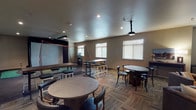 The Golf Room is a well-appointed, single-level recreational space designed to accommodate both golf enthusiasts and social gatherings. At its core is a full indoor golf simulator, complete with a large projection screen, hitting mat, and protective netting, alongside a ceiling-mounted projector for an immersive experience. Adjacent to the simulator area, a compact putting green provides additional practice opportunities on durable flooring.

The lounge portion of the room features patterned carpet tiles in a herringbone-inspired layout, lending a polished, contemporary feel to the space. Comfortable leather seating, including tufted armchairs and sofas, is arranged around a large wall-mounted projection screen, creating a secondary entertainment zone. A mix of round dining tables with chairs and tall bar-height tables with stools offer flexible seating arrangements throughout the room.

Recessed lighting illuminates the space evenly, while multiple windows fitted with white blinds allow for controlled natural light. Neutral taupe walls and a clean, smooth ceiling contribute to the room's cohesive and inviting atmosphere. A dedicated side area with darker flooring houses the simulator bay, clearly delineating the active play zone from the lounge and seating areas.
