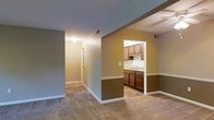 This single-story residence features a practical, open layout that connects the main living area to the kitchen through a distinctive pass-through opening. The living and hallway areas are finished with wall-to-wall carpet in a neutral tone, contributing to a warm and understated ambiance throughout. Walls are painted in a muted olive-green hue with white trim and baseboards, creating a consistent color palette across the space. A two-tone wall treatment, divided by a chair rail, is visible in the kitchen area, adding a subtle architectural detail. The kitchen features medium and dark-toned wood cabinetry on both upper and lower sections, a sink positioned beneath the pass-through, and tile flooring that contrasts with the carpeted areas. Overhead lighting is provided by flush-mounted ceiling fixtures, and HVAC vents are integrated into both the ceiling and walls. A hallway visible from the main living area leads further into the home, suggesting additional rooms beyond. The overall design is straightforward and functional, with a layout that emphasizes connectivity between the primary living and kitchen spaces.