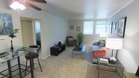 This single-floor residential space offers a comfortable and functional layout designed for everyday living. The living room serves as the heart of the home, featuring soft wall-to-wall carpeting that adds warmth underfoot and a neutral color palette across the walls that creates a clean, inviting atmosphere. A ceiling fan is centrally positioned overhead, providing both air circulation and a practical design element. Natural light flows generously into the room through multiple windows, brightening the space throughout the day. The living area is well-suited for relaxed seating arrangements, as evidenced by its capacity to accommodate a sofa, accent chair, coffee table, and media console with ease. Adjacent to the living room, a dining area is visible and features bar-height seating, suggesting an open-concept connection between the two spaces. A doorway leads further into the home, hinting at additional rooms beyond. The overall ambiance is casual and livable, with a straightforward layout that maximizes usable floor space. The light-toned walls and carpeted floors work together to create a cohesive, understated interior that is both adaptable and welcoming.