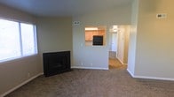 This single-floor space offers a functional and straightforward layout designed for comfortable everyday living. The living area is the central focus, finished with wall-to-wall carpeting in a neutral tone that adds warmth underfoot. A black fireplace anchors one corner of the room, serving as a practical focal point and providing an added layer of coziness to the space. The walls are painted in a soft, muted beige that contributes to a calm and neutral atmosphere throughout.

A pass-through window connects the living area to the kitchen, allowing for an open feel between the two spaces while maintaining a degree of separation. The kitchen features light wood cabinetry and includes a black refrigerator, with overhead lighting that brightens the work area effectively.

From the living room, a carpeted hallway extends toward additional areas of the unit, hinting at further rooms beyond. The hallway is well-lit and features white-painted doors, maintaining the clean and consistent aesthetic seen throughout the space. HVAC vents are integrated into the walls and ceilings, ensuring climate control across the home. Overall, the space presents a clean, move-in-ready environment with a cohesive neutral palette.