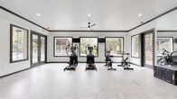 This fitness center is thoughtfully designed across a single level, offering distinct zones that cater to a full range of workout needs. The facility is divided into interconnected spaces, each serving a specific training purpose while maintaining a cohesive modern aesthetic characterized by white walls, dark crown molding, and durable flooring throughout.

The cardio zone is generously proportioned, housing elliptical machines, stationary bikes, stair climbers, and treadmills. Large windows along multiple walls flood the area with natural light and provide views of an outdoor pool and surrounding greenery. Ceiling fans overhead contribute to a comfortable workout environment. Mounted televisions are positioned for easy viewing during cardio sessions.

The strength training area features a motivational accent wall with vertical wood slat paneling and bold "Again & Again" lettering. It is outfitted with Hoist cable machines, a multi-station functional trainer, a dumbbell rack, and various resistance equipment, all set against floor-to-ceiling mirrors that visually expand the space.

A dedicated functional training zone offers open floor space, TRX suspension trainers, resistance bands, plyo boxes, and additional accessories. A compact kitchenette with navy cabinetry, gold hardware, a patterned tile backsplash, and a stainless steel refrigerator completes the facility.