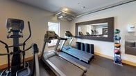 This fitness center offers a practical and well-appointed space for a comprehensive workout experience. The room is finished with durable rubber flooring throughout, providing a safe and resilient surface suitable for a variety of exercises. Exposed metallic ductwork runs along the ceiling, lending an industrial aesthetic that complements the clean white walls and warm wood baseboards.

The cardio zone includes multiple treadmills and an elliptical machine, positioned near large windows that overlook an adjacent courtyard, allowing natural light to fill the space and providing views of the surrounding building exterior. A framed wall mirror reflects the cardio area, enhancing the sense of space.

The strength training area is well-stocked, featuring a multi-function cable machine with cable attachment options, a tiered dumbbell rack holding a full range of free weights, and a flat workout bench. Recessed lighting supplements the natural light, ensuring the space remains well-lit throughout. A glass entry door with keypad access leads directly to the outdoor courtyard, providing convenient exterior access. The overall ambiance is functional and clean, designed to support a full range of fitness activities.