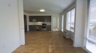 This single-floor space showcases a clean, modern design centered around an open-concept layout that seamlessly blends the kitchen and living areas. The kitchen is outfitted with sleek gray cabinetry, stainless steel appliances including a range, microwave, dishwasher, and refrigerator, all set against a neutral backsplash and light countertops. Warm hardwood-style flooring extends throughout the main living area, adding a sense of warmth and continuity to the space. Large windows with horizontal blinds line one wall, flooding the interior with generous natural light and offering views to the outside. A wall-mounted HVAC unit provides convenient climate control. The open living area offers ample room for a variety of furniture arrangements, making the space highly versatile. A doorway visible in the background suggests access to additional rooms, such as a bedroom or bathroom. The overall ambiance is bright, clean, and contemporary, with a minimalist aesthetic that allows residents to personalize the space to their liking. The combination of modern finishes and an open floor plan creates a functional yet inviting living environment.