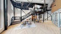 Located at 2940 Falstaff Rd in Raleigh, NC, this striking workspace blends industrial and contemporary design elements into a highly functional and visually compelling environment. The single-floor layout is anchored by soaring double-height ceilings that expose structural steel beams, black metal framework, and industrial ductwork overhead, lending the space an authentic, loft-inspired character. A dramatic black steel staircase with decorative mesh railings serves as a central architectural focal point, leading to an open mezzanine level that overlooks the main floor below.

The main floor features light-toned herringbone hardwood flooring that contrasts beautifully with the dark metal structural elements. Multiple distinct work zones are thoughtfully arranged throughout, including an open collaborative area with a large communal table and mixed seating, a round conference table surrounded by task chairs, and a lounge area with a sofa and wood-topped coffee table. A private office with glazed walls offers a quieter setting with built-in desk, monitor, and curated artwork.

Warm terracotta-toned accent walls add depth and warmth to the open layout, while floor-to-ceiling windows along the perimeter invite abundant natural light and exterior views of the surrounding greenery. Pendant and track lighting supplement the natural light throughout. The overall ambiance is modern, dynamic, and professional.