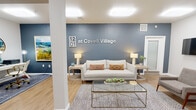 The 1201 at Covell Village leasing office is a single-floor community space designed to offer a polished and inviting first impression. The interior is defined by a cohesive color palette of cool slate blue accent walls contrasted against crisp white surfaces, all anchored by warm-toned light wood flooring that runs throughout the space.

The main lounge area serves as the focal point of the office, featuring a neutral linen sofa accented with patterned throw pillows, flanked by minimalist side tables and lamps. A geometric glass-and-metal coffee table sits atop a textured area rug, giving the seating area a contemporary yet comfortable feel. A wall-mounted flat-screen television adds a functional touch to the lounge.

Adjacent to the lounge is a dedicated workstation area, furnished with a sleek desk, an ergonomic chair, and curated artwork in gilded frames — creating a professional yet approachable workspace. A second private office or consultation room is accessible nearby, appointed with plush seating and patterned carpet, ideal for one-on-one meetings.

The entry corridor features a warm wood credenza topped with decorative accents and a large framed artwork, leading to a glass-paneled door entry. Recessed and flush-mount ceiling lighting throughout ensures the space is bright and well-lit. The overall ambiance is modern, clean, and welcoming — reflective of the community it represents.