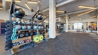 This expansive fitness facility in Denver, Colorado, delivers a well-appointed workout environment within a single open floor plan defined by striking industrial-chic architecture. Soaring ceilings with exposed ductwork, large concrete support columns, and suspended linear lighting create a bold, urban aesthetic throughout the space. Rubber flooring with marked training lanes covers the main workout area, providing both function and visual structure for a variety of training activities.

The facility is comprehensively equipped across distinct zones. A dedicated free weights area houses an extensive selection of kettlebells, medicine balls, dumbbells, and weight plates arranged on organized multi-tier racks. A separate cardio section features rowing machines, air bikes, treadmills, and ski-erg-style equipment. An additional functional training zone incorporates cable machines and specialized strength apparatus. A Jacob's Ladder climber and battle ropes add variety to the training offerings.

A separate studio room with smooth wood-look flooring and warm terracotta accent walls provides a flexible space for stretching, yoga, or group instruction. Large windows throughout invite ample natural light, while full-length mirrors extend the sense of space and support training feedback. Vibrant wall murals with Denver-inspired imagery add a distinctive local character to the overall environment.