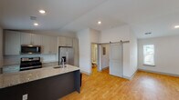 Located in Huntsville, AL (35806), this single-floor space offers a thoughtfully designed interior with modern finishes throughout. The kitchen serves as a central highlight, featuring a large island with granite countertops, a deep sink, and a full suite of stainless steel appliances including a gas range, built-in microwave, French door refrigerator with ice maker, and dishwasher. Crisp white upper cabinetry pairs with darker lower cabinets for a contemporary contrast, while recessed lighting and vaulted ceiling details add a sense of openness. Warm-toned hardwood flooring flows seamlessly throughout the main living areas, complementing the bright, neutral wall palette. A stylish sliding barn door adds a distinctive architectural touch, providing access to the bathroom. The bathroom is clean and modern, equipped with a granite-topped vanity, framed mirror, walk-in glass-enclosed shower, and wood-toned flooring. The open living area benefits from natural light through multiple windows with blinds. A carpeted stairwell with dual handrails provides access to additional levels. Overall, the space presents a clean, contemporary aesthetic with quality finishes and functional design.