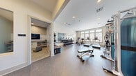 Located in Spring, TX, this fitness center offers a comprehensive exercise facility designed to support a wide range of workout routines. The space is laid out on a single floor and is organized into distinct functional zones that flow seamlessly together.

The main exercise area features durable rubber flooring and is filled with an extensive selection of equipment, including multiple treadmills, an elliptical machine, a stationary bike, and a rowing machine positioned along large, multi-pane windows that allow ample natural light and outdoor views of the surrounding community. A full rack of dumbbells lines one wall, accompanied by a large mirror that enhances the sense of openness. A functional cable machine and adjustable weight bench provide options for strength training, while a plyo box and wall-mounted exercise mat support functional and bodyweight workouts.

Recessed lighting throughout maintains a clean, well-lit environment. A built-in water fountain station is conveniently located near the entrance, alongside access to an adjacent lounge area furnished with a wall-mounted television, seating, and cabinetry — ideal for pre- or post-workout relaxation. The neutral color palette of whites and grays, combined with the bright, open layout, contributes to a modern and motivating atmosphere.