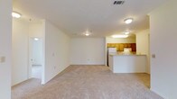 This single-floor residence offers a clean, open-concept layout that seamlessly connects the living area to the kitchen. The spacious living room is finished with plush, light-toned carpeting underfoot and bright white walls that lend a fresh, airy feel to the space. Flush-mounted ceiling lights provide consistent illumination throughout. The kitchen is positioned at the far end of the open floor plan and is defined by warm honey-toned wood cabinetry that extends along both the upper and lower sections, offering ample storage. A white refrigerator and a peninsula-style breakfast bar anchor the kitchen, with the bar serving as a natural divider between the cooking and living spaces. The peninsula provides a functional surface ideal for casual dining or food preparation. A built-in overhead light fixture above the kitchen area ensures the workspace is well-lit. HVAC vents are integrated into the ceiling, maintaining comfort throughout the home. The overall ambiance of the space is bright, practical, and welcoming, with a neutral color palette that creates a versatile backdrop suited to a variety of interior styles. The open flow of the floor plan maximizes the sense of space and promotes easy movement between living and cooking areas.