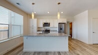 Located at 210 W Fifth Ave in Columbus, Ohio, this single-floor unit offers a thoughtfully designed open-concept layout that seamlessly connects the kitchen, dining, and living areas. The kitchen serves as the centerpiece, showcasing a large granite-topped island with an undermount sink, white shaker-style cabinetry, and a full suite of stainless steel appliances including a French door refrigerator, electric range, and built-in microwave. Pendant lighting above the island adds a modern touch, while wood-look plank flooring extends throughout the main living area. The living space is bright and airy, featuring French doors that open to a private balcony and allow generous natural light to fill the room. A stylish barn door leads to the bedroom wing, adding a contemporary architectural detail. The primary bedroom features plush carpeting, a large window with outdoor views, and access to a generously sized walk-in closet outfitted with wire shelving. The bathroom is well-appointed with a dual-sink granite vanity, a glass-enclosed walk-in shower with built-in shelving, and wood-look flooring. An additional carpeted walk-in closet and a dedicated in-unit laundry space with a stacked washer and dryer round out the practical amenities of this well-equipped residence.
