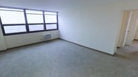 This single-floor space presents a clean and functional layout well-suited for a variety of uses. The main room is generously proportioned, featuring neutral wall-to-wall carpeting that adds a sense of warmth underfoot while complementing the room's simple, understated aesthetic. The walls are finished in a smooth, bright white, contributing to an open and airy atmosphere throughout.

A prominent set of wide, horizontal windows spans one corner of the room, allowing ample natural light to filter in and offering views of the surrounding urban environment. The windows are fitted with roller blinds, providing flexible light and privacy control. Beneath the windows sits a through-wall air conditioning unit, ensuring climate comfort during warmer months.

The room is defined by its simplicity and versatility — the open floor plan and absence of built-in fixtures make it highly adaptable to different configurations and purposes. Dark baseboard trim runs along the perimeter of the room, providing a clean visual contrast against the light-colored walls and carpet. Electrical outlets are positioned along the walls, offering practical access points throughout the space. Overall, the space conveys a straightforward, functional character with a neutral palette ready to be personalized.