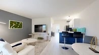 This thoughtfully designed single-floor unit offers a seamless open-concept layout that maximizes every square foot of its compact footprint. The combined kitchen and living room serves as the heart of the home, where a striking kitchen takes center stage with two-tone cabinetry — bold navy lower cabinets paired with crisp white upper cabinets, all accented by brushed gold hardware. Marble-look countertops and a matching backsplash add a luxurious touch, complemented by stainless steel appliances and a gold-finish faucet. A peninsula with bar seating flows naturally into the dining and living areas, creating an ideal layout for entertaining. The living space features light wood-look flooring, a marble-surround media console, and a charcoal accent wall with decorative greenery art, adding depth and character to the open room. Natural light enters through vertical-blind windows. The spacious bedroom offers a wide mirrored sliding-door closet, a large window overlooking greenery, and a clean, neutral palette. The hallway connects to the full bathroom, which includes a tiled shower-tub combo, light wood-look flooring, and gold accent fixtures, maintaining a cohesive modern aesthetic throughout.