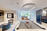 This expansive game room in Alexandria, Virginia, is a thoughtfully designed recreational facility that blends modern aesthetics with a diverse range of entertainment options across a single, well-appointed floor. The space is divided into distinct zones that flow seamlessly into one another, creating an engaging and versatile environment.

At the heart of the main lounge area sits a sleek, contemporary billiards table anchored by a bold blue patchwork area rug. Adjacent to it, a shuffleboard table is framed by a built-in wood-paneled entertainment wall with display shelving and an overhead TV. Seating vignettes with modern accent chairs and ottomans are arranged throughout for casual gathering.

A separate lounge zone features deep navy walls adorned with large-scale artwork, a curved sectional sofa, and an eye-catching sculptural ceiling installation with globe pendant lights. A built-in wet bar with warm wood cabinetry, a large-screen TV, and open shelving provides an ideal setting for social occasions.

The facility also includes a full-size hardwood sports court with padded navy wall panels, high ceilings, interior viewing windows, and inspirational typographic murals. Floor-to-ceiling windows in the main lounge area draw in natural light and connect the interior to outdoor greenery, enhancing the overall ambiance of this premier recreational space.